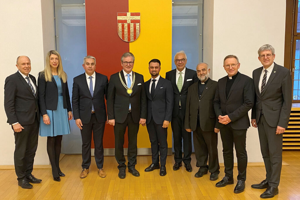 Acht Männer in Anzügen und eine blonde Frau im blauen Kleid stehen vor einer rot-gleben Wand mit dem Wappen von Paderborn. Es sind die Vertreter der Religionsgemeinschaften, der Landrat des Kreises Paderborn und der Bürgermeister mit gloldener Kette